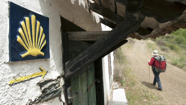 A hiker walks along a dirt path near a rustic house marked with a scallop shell symbol and directional arrow for pilgrims.