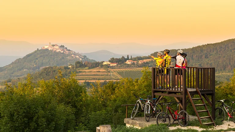 Two cyclists in bright gear admire the view from a wooden platform, overlooking vineyards and a hilltop village at sunset.