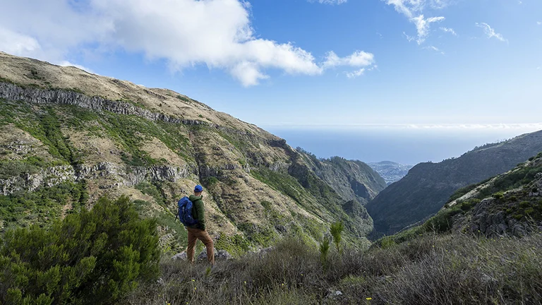 A hiker stands on a rocky hillside, gazing at the lush green valleys and blue ocean under a cloudy sky.