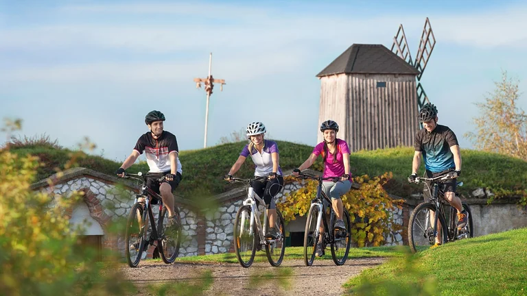 Four cyclists poised on mountain bikes against a scenic background featuring a traditional windmill and foliage.