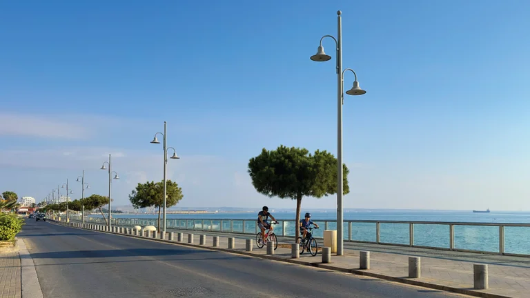 Two cyclists pause on a seaside promenade lined with lamps and palm trees, overlooking a calm blue sea under a clear sky.