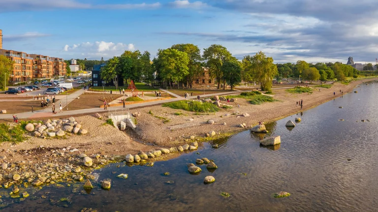 A scenic waterfront view with people strolling along a sandy beach, surrounded by trees and modern buildings under a partly cloudy sky.
