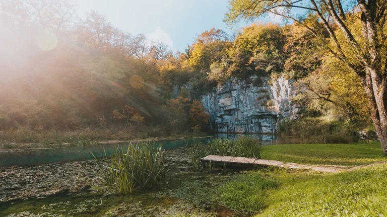 A serene lake surrounded by autumn foliage and a rocky cliff, with a wooden dock extending into the water under a bright sky.