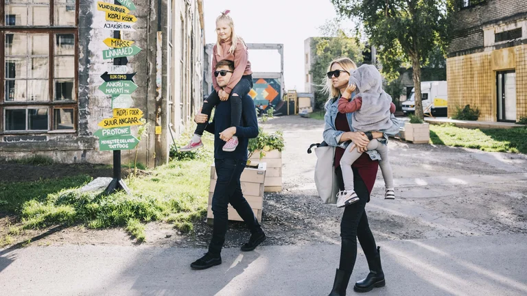 A family walks together outdoors, with children on shoulders, beside a signpost displaying colorful directional signs.