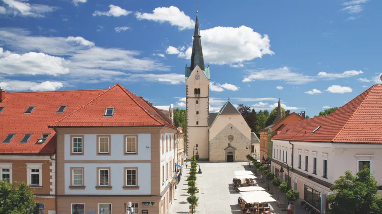 A sunny town square featuring historic buildings with red roofs and a tall church steeple against a backdrop of blue skies and clouds.