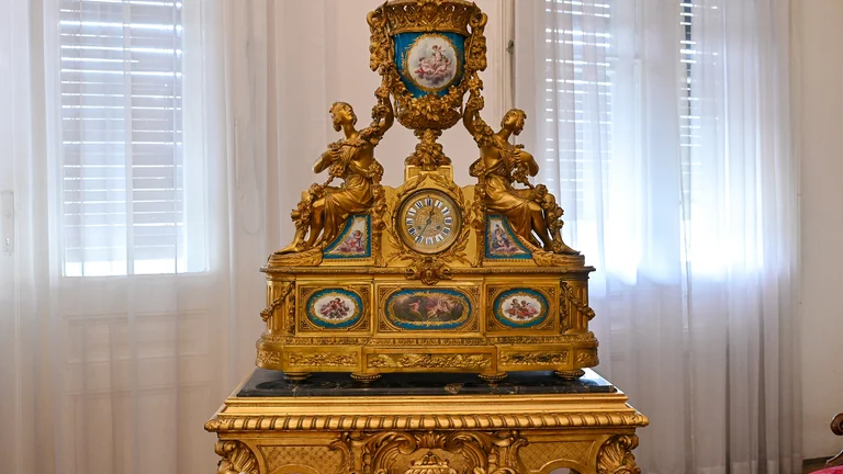 An ornate golden clock adorned with figurines and paintings sits atop a lavish table, illuminated by soft natural light filtering through sheer curtains.