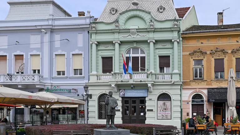 A photograph of a historic green building with flags, surrounded by pastel-colored structures and outdoor dining areas.