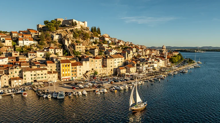 A picturesque coastal town with colorful buildings and a marina, overlooked by a medieval castle on a hill, bathed in golden sunlight.