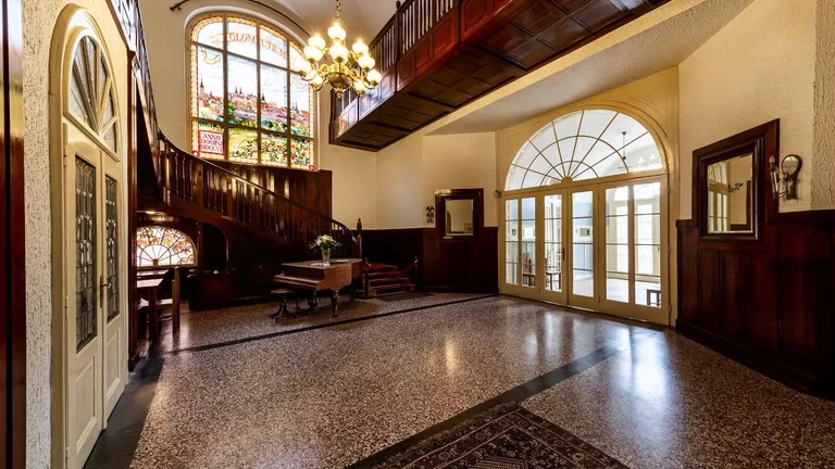 A spacious, elegant lobby featuring a grand staircase, stained glass windows, a vintage piano, and intricate wood paneling.