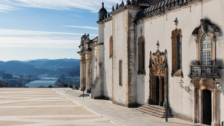 A historic building with ornate architecture overlooks a scenic view of hills and a river under a clear blue sky.