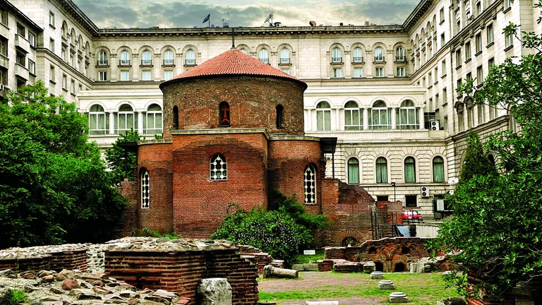Ancient red-brick Rotunda of St. George surrounded by modern buildings, with visible Roman ruins in the foreground.