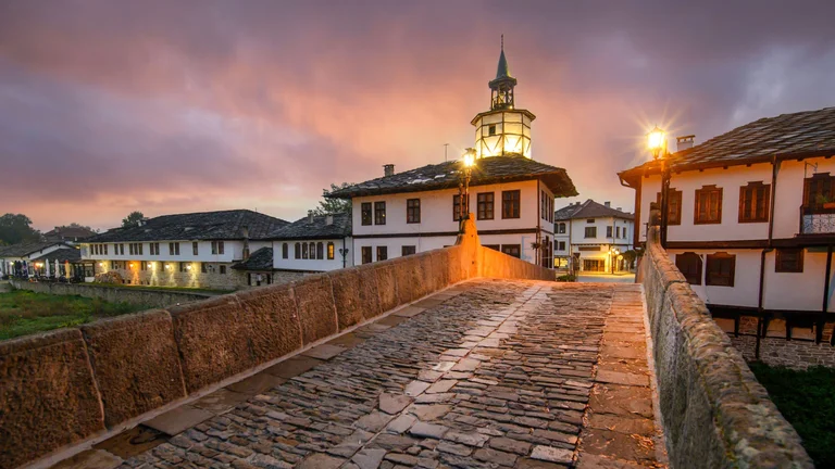 Historic cobblestone bridge leading to the illuminated Clock Tower in the old town of Tryavna at dusk.