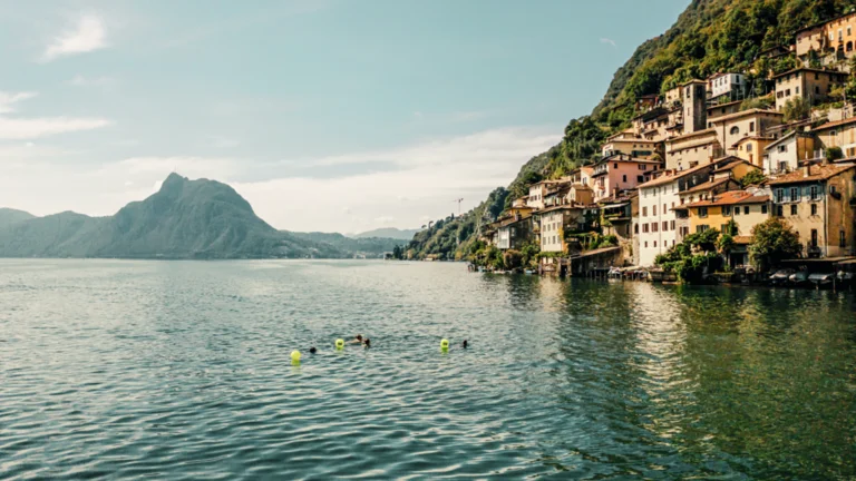 Serene lakeside scene with swimmers and colorful buoys, surrounded by picturesque hills and charming village buildings under a clear sky.