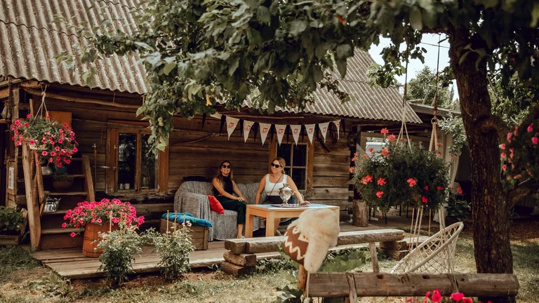 Two women relaxing on the porch of a rustic wooden house on the Onion Route by Lake Peipsi, Estonia, decorated with flowers and traditional details.