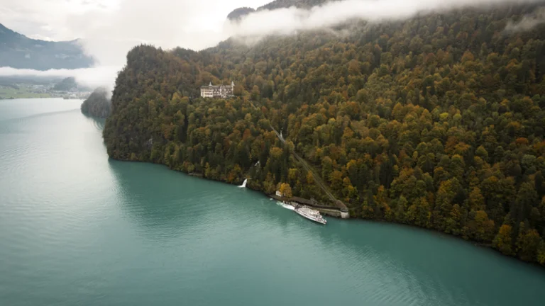 A serene lake surrounded by autumn-colored forests and misty mountains, with a historic building perched on a cliff and a boat at the shore.