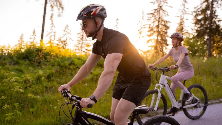 Two cyclists riding electric mountain bikes along a forest path at sunset, with golden light filtering through trees.