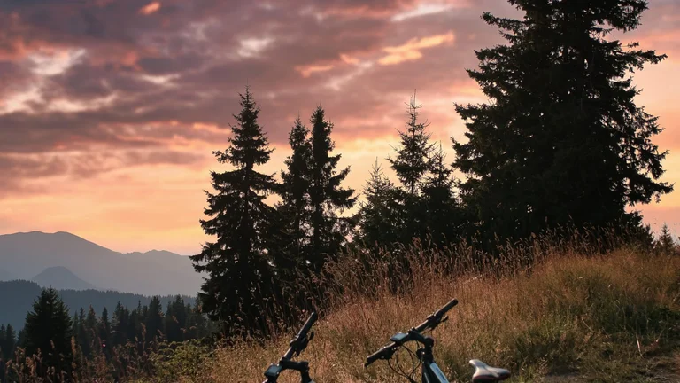 Two electric bikes parked on a grassy hilltop trail, with a dramatic sunset and mountain silhouettes in the background.