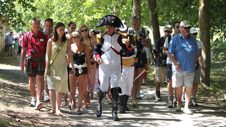 A group of people walks along a path, with one individual dressed in a historical military uniform, surrounded by spectators.