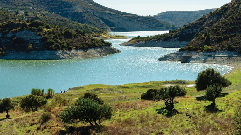 A peaceful view of Germasogeia Dam with winding turquoise waters, green hills, and scattered olive trees.