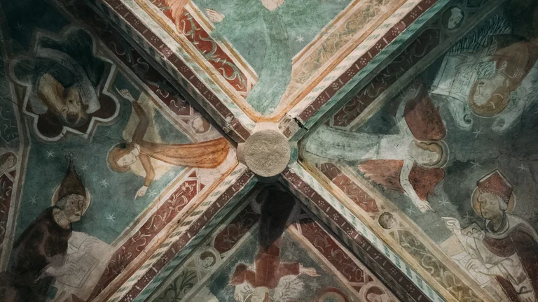 This picture shows an old, painted vaulted ceiling with religious frescoes in muted colors, featuring figures like saints or evangelists against a starry, teal-green background. The ceiling ribs form an 'X' pattern, meeting at a central circular boss.