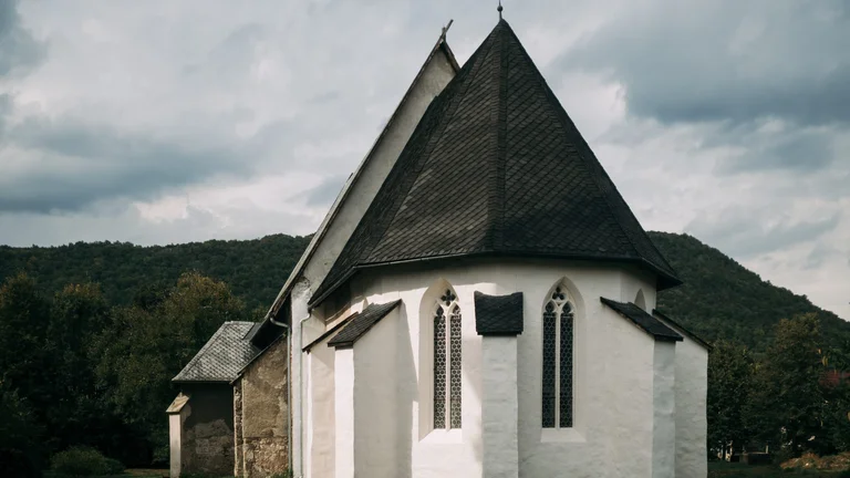 Small white Gothic church of Plešivec in Slovakia on a cloudy day, with a pointed roof and surrounded by forest, sunlight casting soft shadows on its exterior walls.