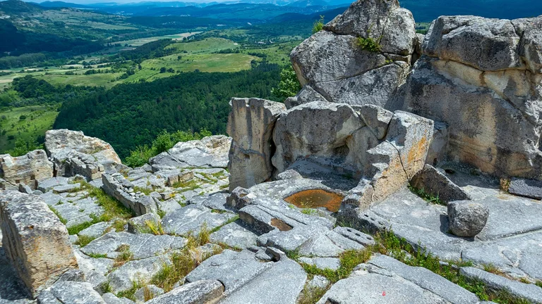 Aerial view of the ancient Thracian city of Perperikon in Bulgaria, with rock-hewn ruins atop a hill and mountains visible in the background surrounded by green terrain.