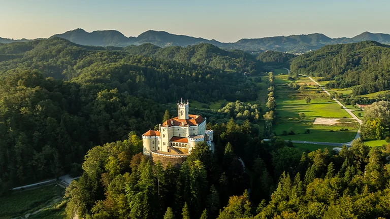 Aerial view of Trakošćan Castle, a white and red-roofed historic fortress surrounded by dense green forest and rolling hills in northern Croatia. The castle sits atop a wooded hill, overlooking a lush valley and a winding country road under a clear blue sky.