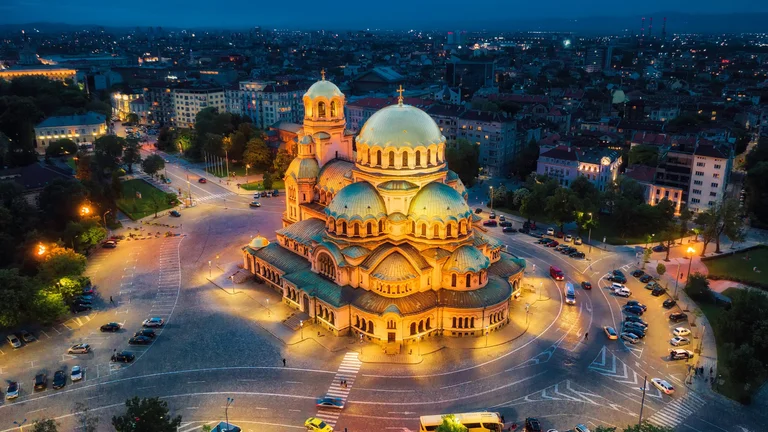 Vue aérienne nocturne de la cathédrale Alexandre Nevski à Sofia, en Bulgarie, illuminée d’une lumière dorée sur fond de paysage urbain sombre.