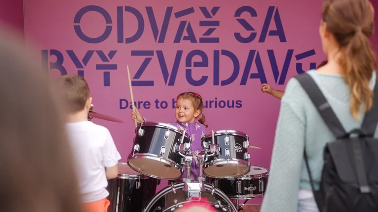 Une jeune fille joue de la batterie avec enthousiasme devant une pancarte rose sur laquelle est écrit « Osez être curieux » lors d’un événement en plein air.