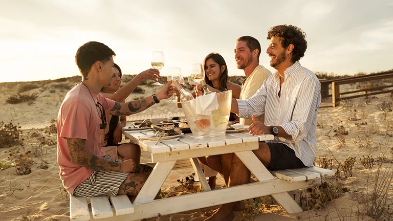 晴れた日に、ビーチの木製ベンチテーブルに座りながら、友人たちが笑顔でワイングラスを掲げて乾杯している。