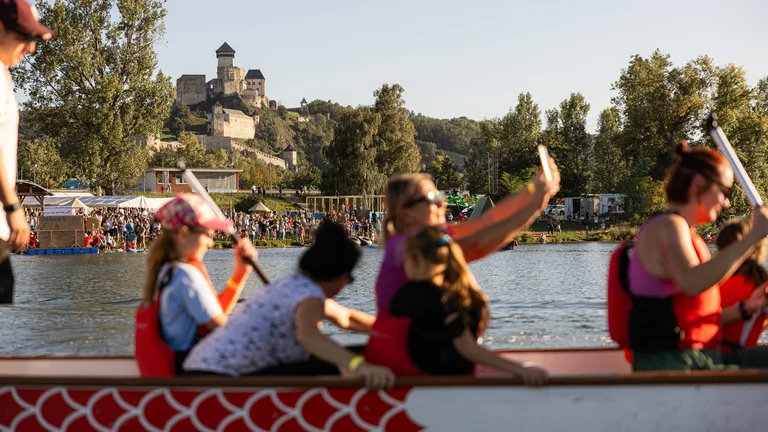 Course de bateaux-dragons avec des personnes pagayant sur une rivière, le château de Trenčín étant visible sur une colline boisée en arrière-plan.