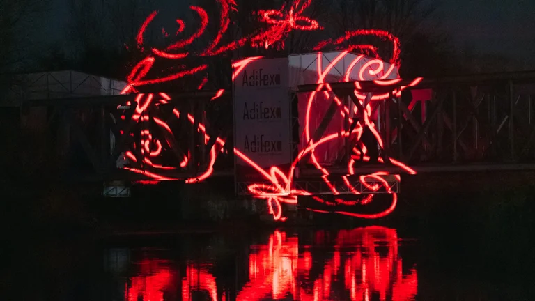 Traînée lumineuse rouge en forme de scooter se reflétant clairement sur l’eau sombre lors d’un événement nocturne de lumières.