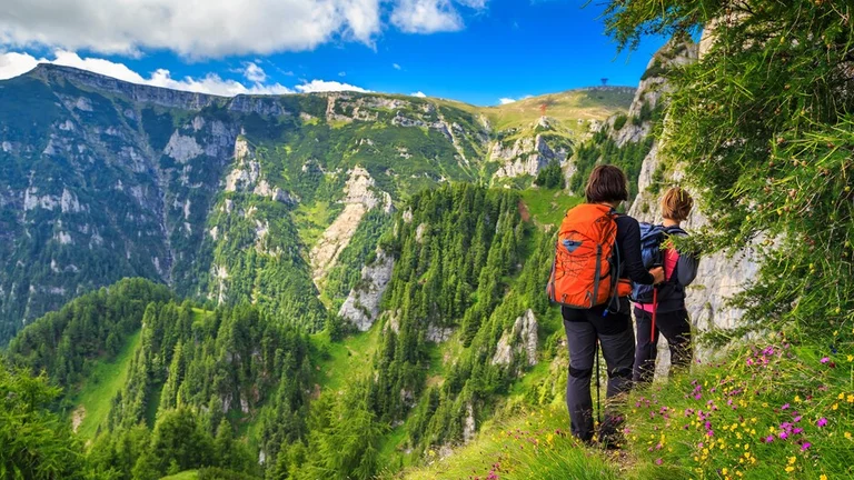 Two hikers with backpacks standing on a mountain trail overlooking a deep green valley in the Carpathian Mountains.