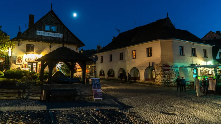 Historic town square at night with illuminated arcaded buildings and a bright moon overhead