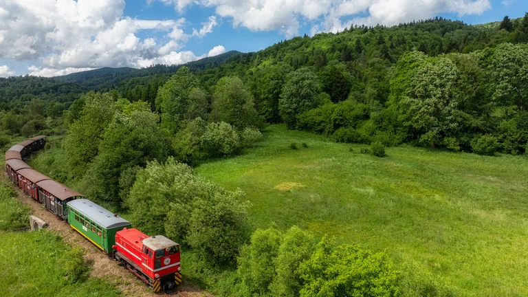 Red and green train traveling through lush green countryside under a partly cloudy sky.