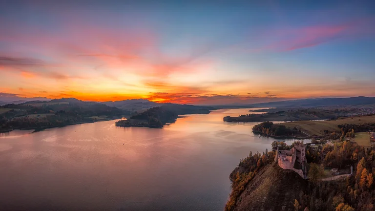 Castle overlooking a river at sunset with vibrant sky and distant hills.
