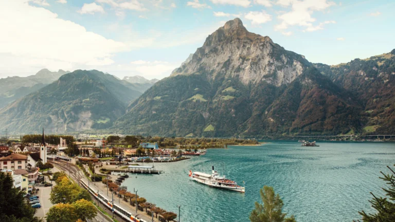 Train and boat by a lakeside village, with mountains in the background under a cloudy sky.