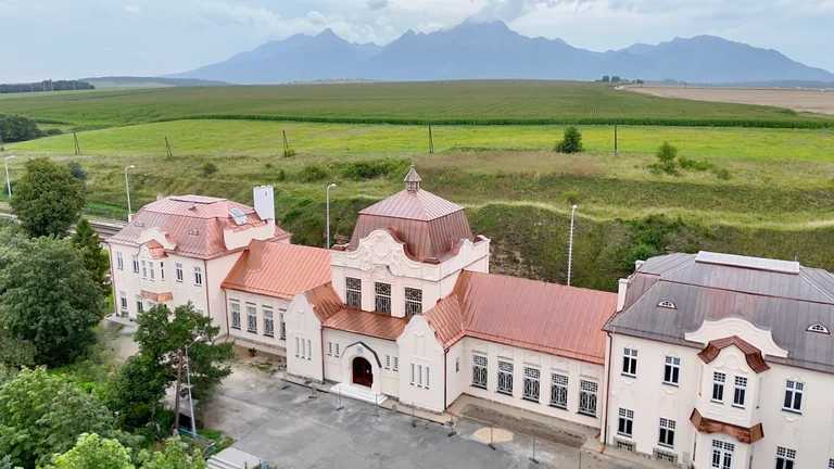 Large historic building with red roofs, fields behind, and mountains in the distance.