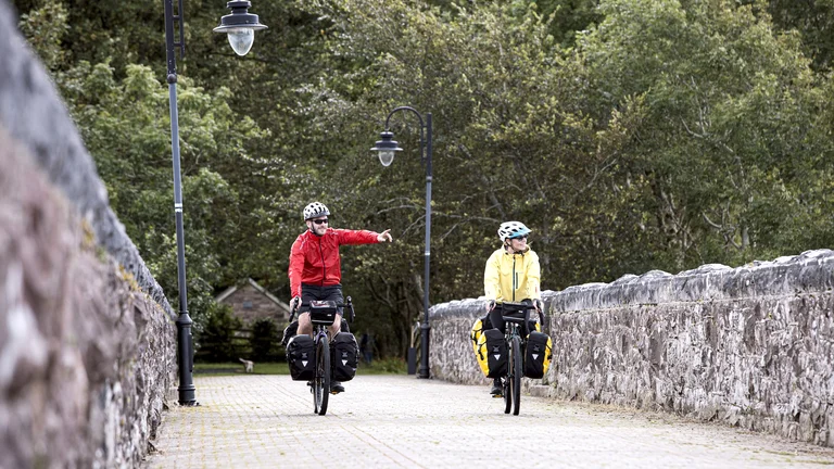 Two cyclists with gear ride on a stone bridge surrounded by trees.