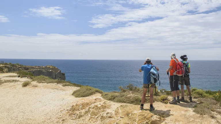 Three hikers with backpacks stand on a cliff overlooking the ocean under a blue sky.