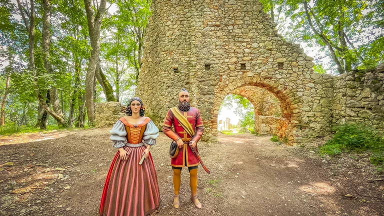 Two people in medieval costumes stand before a stone ruin in a wooded area.