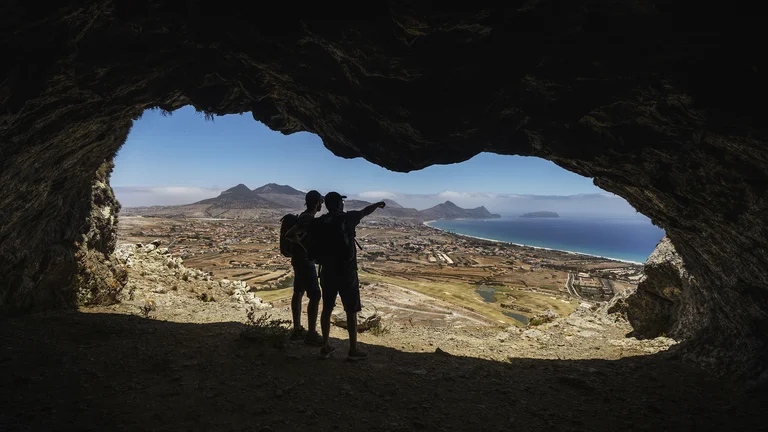 Two people stand in a cave, looking out at a coastal landscape under a clear sky.