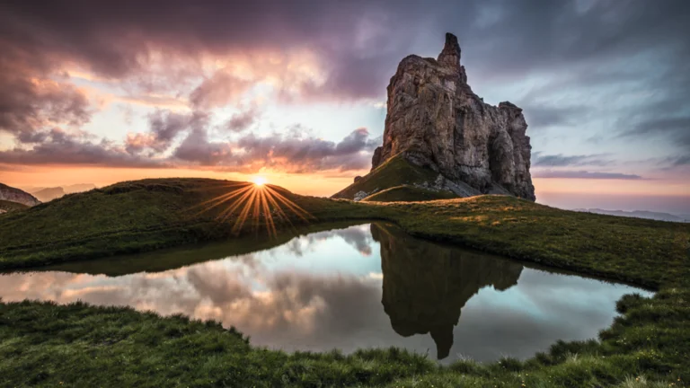 Rocky hill and dramatic sunset reflected in a calm pond, surrounded by green grass.