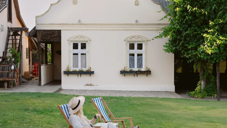Person relaxing on a lawn chair with a book in front of a quaint house on a sunny day.