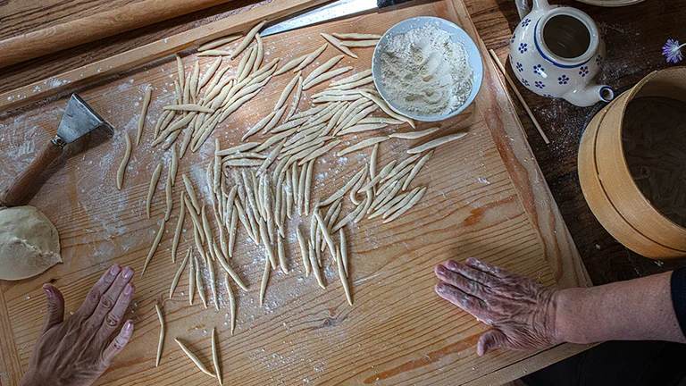 Hands shaping fresh pasta on a floured wooden board with dough and tools nearby.