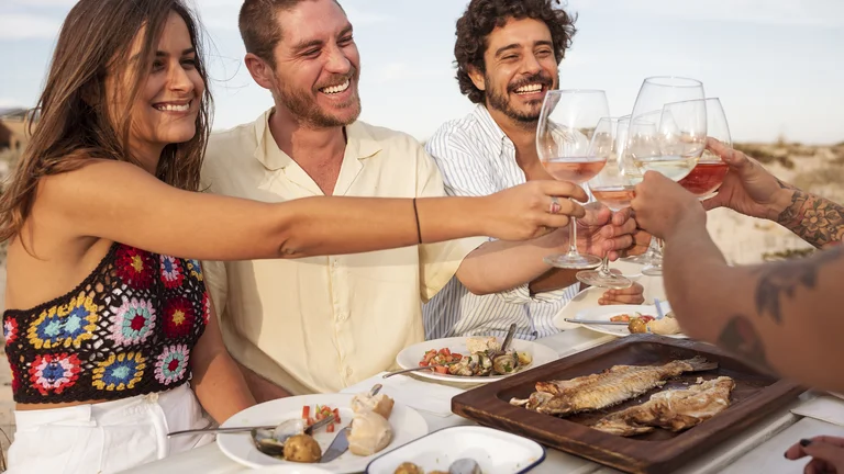 Friends cheerfully clinking wine glasses around a table with grilled fish outdoors.