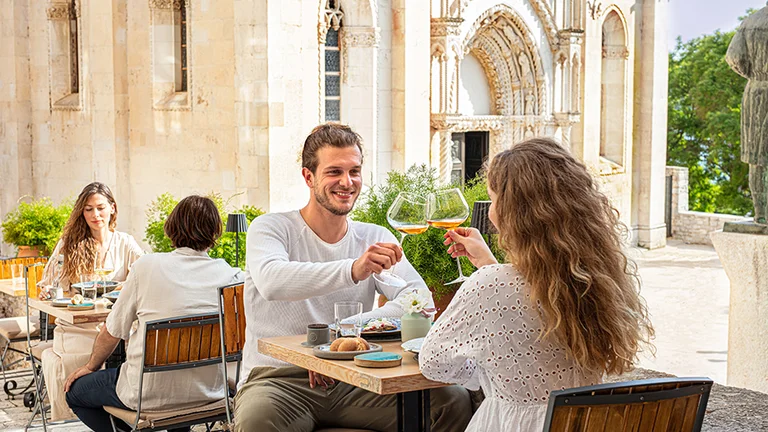 Smiling couple clinking glasses at an outdoor café near a historic building.