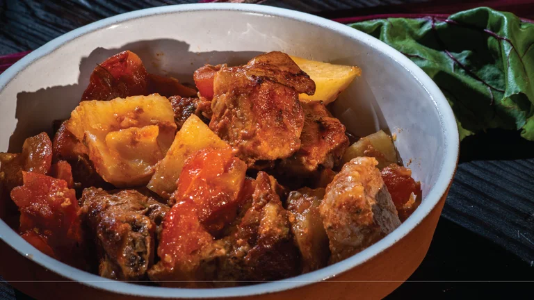Bowl of stew with chunks of meat, tomatoes, and yellow vegetables on a dark table.