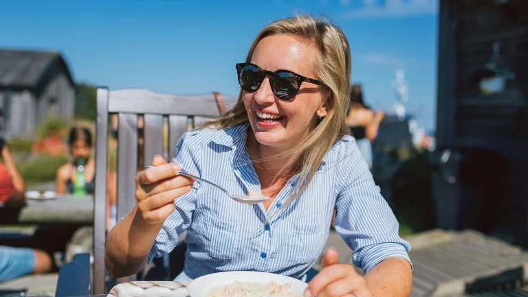 Smiling woman in sunglasses eating soup at an outdoor restaurant on a sunny day.