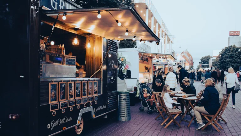 People dining at outdoor tables near food trucks on a city street in the evening.